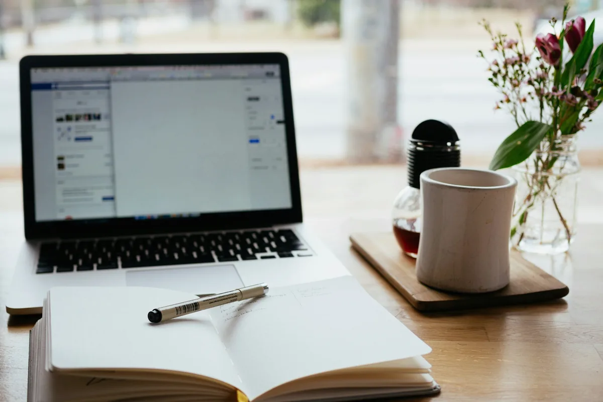 Table près d'une fenêtre : ordinateur portable ouvert, carnet, stylo et petit bouquet